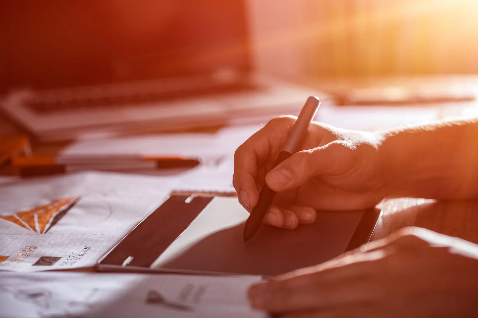 Graphic designer using a digital pen tablet to sketch logo concepts and brand visuals beside paper proofs and a laptop in a sunlit creative workspace.